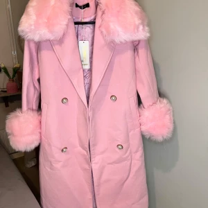 Rosa kappa med fuskpäls och pärlknappar med bälte  - Supersöt rosa kappa med fluffig fuskpäls på krage och ärmslut. Dubbelknäppt med stora pärlknappar och quiltat foder på insidan. Perfekt statement-plagg för dig som vill sticka ut med en gullig och trendig look.