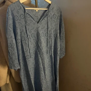 Blå broderad klänning från Indiska - Säljer en ljusblå klänning från Indiska i storlek M. Klänningen har v-ringning med knytband, trekvartsärm och är täckt av broderade blommor med hålmönster. Materialet känns lätt och luftigt, perfekt för varmare dagar.