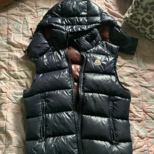 Mörkblå dunväst från Moncler - Snygg mörkblå dunväst från Moncler med avtagbar huva och brunt foder. Västen har två dragkedjefickor, quiltad design och klassisk Moncler-logga på bröstet. Insidan har ett coolt serietryck och stor Moncler-etikett. Perfekt för kyliga dagar. Lite skadad i luvan där man knäpper