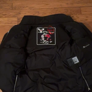 Svart dunjacka Moose Knuckles - Svart dunjacka från Moose Knuckles med coolt broderat märke på insidan. Jackan har dragkedja, stora fickor med tryckknappar och detaljer i silver. Perfekt för kalla dagar och har en modern, sportig look.
