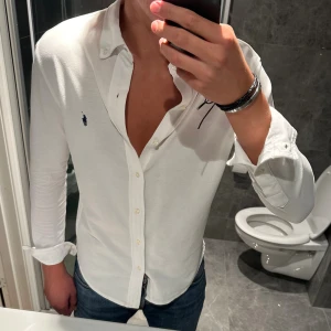 Vit skjorta från Ralph Lauren - Klassisk vit skjorta från Ralph Lauren med blå broderad logga på bröstet. Skjortan har lång ärm, knappar hela vägen och en snygg krage. Tillverkad i mjuk bomull och har en normal passform som sitter snyggt på kroppen. Skjortan är i riktig bra skick! Jag är 188cm och bär storlek S och den är något tight på mig. Hör av er vid frågor;)