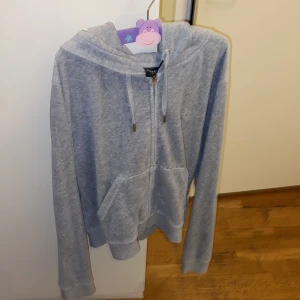 Grå hoodie från Juicy Couture - Grå hoodie från Juicy Couture med dragkedja, snörning i huvan och broderad logga på bröstet. Mjuk och skön med känguruficka framtill och långa ärmar. Perfekt för chill dagar eller när du vill ha en avslappnad look.