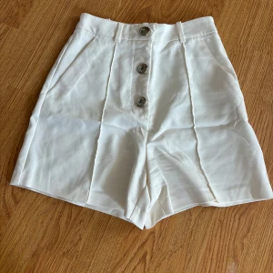 Vita högmidjade shorts från Zara - Snygga vita shorts från Zara med hög midja och tre silvriga knappar framtill. De har en klassisk, rak passform och är tillverkade i ett mjukt bomullsmaterial. Shortsens stilrena look och bälteshällor gör dem perfekta för en fräsch sommaroutfit.