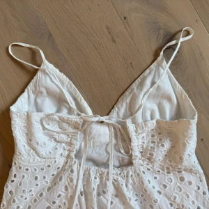 Vit långklänning med broderade detaljer - Superfin vit långklänning med tunna axelband och v-ringning. Klänningen har broderade hålmönster och volang nertill, samt knytning i ryggen. Perfekt för sommarens alla tillfällen och ger en romantisk vibe.