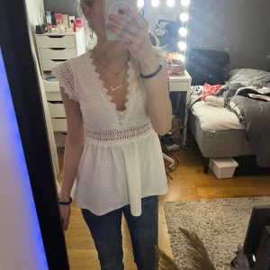 Vit spetsblus med v-ringning - Superfin vit blus med djup v-ringning, spetsdetaljer och broderade hålmönster. Blusen har korta volangärmar och markerad midja med bred spets. Perfekt till jeans för en söt och somrig look.