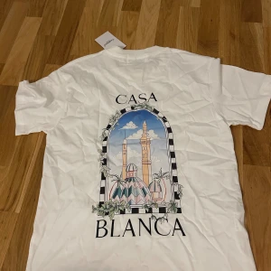 Vit t-shirt från Casablanca med tryck - Vit t-shirt från Casablanca med färgglatt grafiskt tryck av byggnader och växter på ryggen samt mindre tryck på bröstet. Klassisk passform, rund hals och korta ärmar. Perfekt för dig som gillar stilrena men unika plagg.