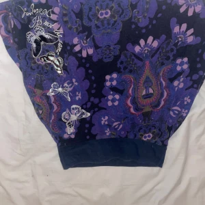 Lila blommig kjol med fjärilar Desigual - Unik kjol från Desigual i lila och mörkblå toner med blommigt mönster och broderade fjärilar. Tröjan har bred mudd nertill och coola detaljer med text och brodyr. Perfekt för dig som gillar färg och mönster! ( bilden råka bli upp och ned)