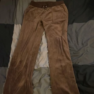 Bruna velourbyxor Juicy Couture S - Mysiga bruna mjukisbyxor från Juicy Couture i velour med snörning i midjan och två stora fickor fram. Klassisk broderad JC-logga på ena fickan och bred resår i midjan. Perfekta för chill eller streetstyle.