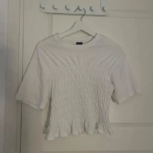 Vit smockad blus med volangkant - Supersöt vit blus med smockad midja och volangkant nertill. Gjord i mjukt bomullstyg som känns skönt mot huden.