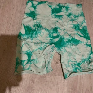 Grön tie-dye träning shorts - Snygga högmidjade shorts med grön och vit tie-dye mönster. De är stretchiga och har en scrunch-detalj bak för extra passform. Perfekta för gymmet eller andra sportaktiviteter. Materialet känns som mjuk och följsam syntet, vilket gör dem bekväma att röra sig i.