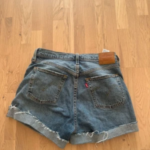 Levi's blå jeansshorts med rå kant - Säljer ett par klassiska blå jeansshorts från Levi's med hög midja och rå, fransig kant. Shortsen har fem fickor, knappgylf och den ikoniska Levi's-lappen bak. Perfekt för dig som gillar en chill och avslappnad stil.