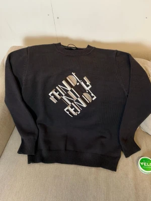 Svart stickad tröja från Fendi - Svart stickad tröja från Fendi med rund hals och ribbade muddar. Framsidan har ett grafiskt Fendi-tryck i vitt och beige som ger en cool vibe. Perfekt för dig som gillar designer och vill sticka ut med en snygg logga på bröstet. Obs säljs endast på https://modeza.se