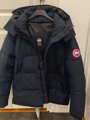 Mörkblå dunjacka från Canada Goose - Säljer en mörkblå dunjacka från Canada Goose med huva och klassisk logga på ärmen. Jackan har quiltad design, dragkedja och knappar framtill. Perfekt för kalla dagar och har en snygg, normal passform som funkar till de flesta stilar.