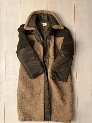 Beige och grön teddyjacka från Kappahl - Säljer en lång teddyjacka från Kappahl i beige och mörkgrön färg. Jackan har quiltad insida, höga krage, dragkedja och detaljer i polyester på ärmar och fickor. Perfekt för kalla dagar och har en loose passform.