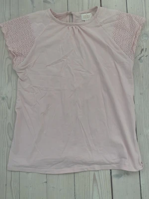 Ljusrosa blus från Creamie - Säljer en ljusrosa blus från Creamie i storlek 152. Toppen har korta ärmar med broderad spets och vågigt mönster, samt rund halsringning och knappöppning bak. Materialet är mjukt och lätt bomull, perfekt för vår och sommar.