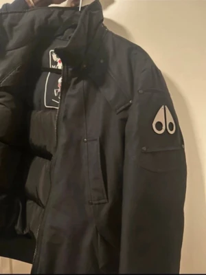 Svart dunjacka från Moose Knuckles - Nyskick: 10000kr. Säljer pga kommer inte till användning. Snygg svart dunjacka från Moose Knuckles med hög krage, ribbade muddar och dragkedja framtill. Jackan har flera fickor med knappdetaljer och ett stort broderat Moose Knuckles-märke på insidan. Perfekt för kalla dagar och har en cool streetstil.