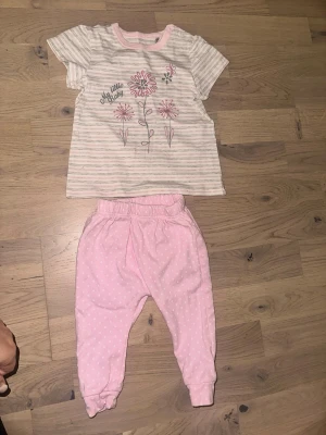 Fint Klädset - T-shirt och Byxor Randig babyväst från Kanz - Ett sött set bestående av en t-shirt med blommotiv och matchande byxor med prickar. Säljer en söt babyväst från Kanz i ljusrosa och vit med tunna ränder och blommigt tryck framtill. Västen har rund hals med tryckknappar på axeln och är gjord i mjuk bomull. Perfekt för små barn som gillar gulliga detaljer och mjuka material.