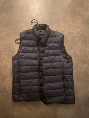 Mörkblå dunväst från Uniqlo - Säljer en mörkblå, quiltad dunväst från Uniqlo i modellen Ultra Light Down. Västen är lätt, har hög krage och är tillverkad i syntetmaterial med vattenavvisande yta. Perfekt att slänga över en hoodie eller tröja för extra värme. Sälj pga för liten på mig☺️ (BILLIGARE VID SNABB AFFÄR)