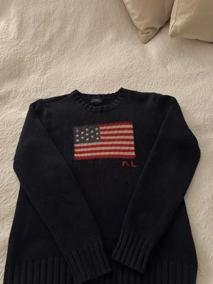 Ralph Lauren USA flagga tröja - Säljer en riktigt schysst och populär stickad tröja från Ralph Lauren. Tröjan är mörkblå och är väldigt tjock och varm vilket passar bra nu till vintern. Storleken är 14-16 vilket är ungefär Xs i vuxenstorlek och passar dig mellan 160-170 cm. Skicket är 9/10 då den knappast är använd. Skriv vid ytterligare frågor!✌️