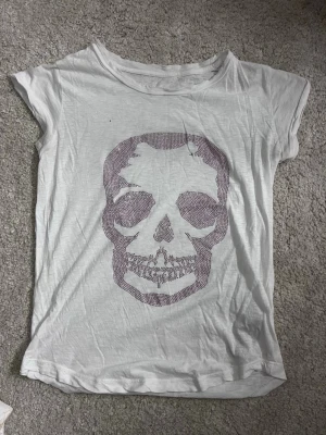 Vit t-shirt med döskalle från Zadig & Voltaire - Vit t-shirt från Zadig & Voltaire med korta ärmar och ett stort döskalletryck i glittriga stenar framtill. T-shirten är i mjuk bomull och har en avslappnad passform. Perfekt för dig som gillar edgy detaljer och vill sticka ut lite extra.