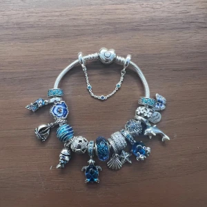 Pandora armband i silver med blå/hav berlocker - Havsinspirerat Pandora armband i silver med massor av blå och silverfärgade berlocker. Den har en säkerhetskedja och två klämstopp som ser till att berlockerna inte trillar av armbandet. Så gott som nytt (använt färre än 10 gånger) och det finns inga defekter. 20% billigare än riktiga priset. Alla delar kan köpas separat (se andra annonser).                                                                    *Obs! Papegojan och delfinen är sålda.