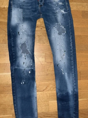 Dsquared2 blå jeans med slitningar - Säljer ett par blå jeans från Dsquared2 med coola slitningar och färgstänk på benen. Jeansen har klassisk femficksdesign, knappgylf och tydliga logodetaljer både fram och bak. Passformen är smal och materialet är robust denim i bomull.
