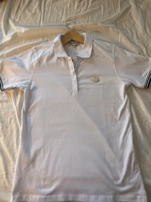 Vit Moncler pikétröja XL - Säljer en stilren vit pikétröja från Moncler i storlek XL. Tröjan har klassisk krage, knappslå med tre knappar och Moncler-logga på bröstet. Ärmarna är korta med diskreta ränder vid mudden. Perfekt för dig som gillar exklusiva märken och enkel design. Pass formen är 40 i axelbredd och 65 längd så passar Storlek S och M. Pris går att diskutera 