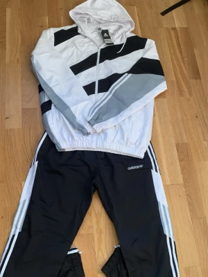 Adidas vindjacka & byxor set – svart/vitt/grått - Sportigt och stilrent Adidas-set bestående av lätt vindjacka med huva och matchande byxor. Design i svart, vitt och grått med klassiska Adidas-ränder och logotyper. Perfekt för träning, promenader eller vardagsbruk. Bekväm passform och lätt material som passar året runt. Ett komplett set med modern look.