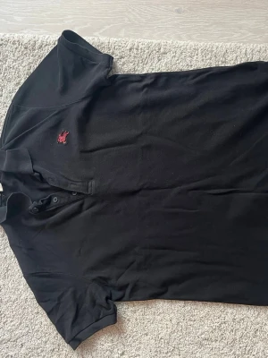 Svart Burberry piké med röd logga - Säljer en svart pikétröja från Burberry i storlek L. Tröjan har korta ärmar, klassisk krage och knäppning med tre svarta knappar. På bröstet finns Burberrys röda broderade logga. Materialet är mjuk bomull som känns skönt mot huden. Självklart är den äkta