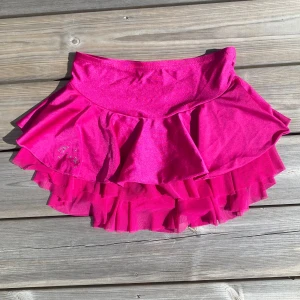 Konståknings volangkjol i stark rosa - Supersöt kort volangkjol i rosa med glittriga stenar på ena sidan. Kjolen har flera lager med volanger och är gjord i ett glansigt, stretchigt material som ger en riktigt poppig look. 😇