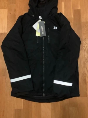 Svart vindjacka från 151FUNK med huva - Svart vindjacka från 151FUNK med huva och reflekterande detaljer på ärmar och rygg. Jackan är vatten- och vindtät med dragkedja framtill och två bröstfickor. Perfekt för blåsiga och regniga dagar, tillverkad i slitstark polyester.