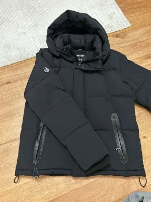 Svart pufferjacka från Superdry - Svart pufferjacka från Superdry med huva och stora, snygga dragkedjor på fickorna. Jackan har en quiltad look och är perfekt för kalla dagar. Märket syns tydligt på insidan och på ärmen. Passformen är normal och jackan har långa ärmar.