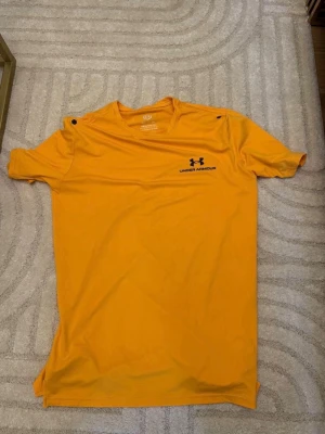Gul t-shirt från Under Armour - Säljer en gul t-shirt från Under Armour med svart logga och text på bröstet. T-shirten har rund hals och korta ärmar, perfekt för träning eller chill. Materialet är lätt och känns skönt mot huden. Snygg och sportig stil!