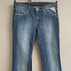 Blå bootcut jeans från Replay 26/34 - Snygga blå jeans från Replay med bootcut passform och klassisk femficksdesign. Jeansen har ljusa slitningar framtill, broderade detaljer på bakfickorna och en rosa patch med blommor bak. Tillverkade i jeansmaterial med låg midja. Midjemått ca  70 cm kanske ngn cm mer