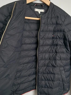 Svart pufferjacka Tommy Hilfiger XS - Snygg svart pufferjacka från Tommy Hilfiger i storlek XS. Jackan har quiltad design, dragkedja framtill och diskret logga på bröstet. Fodrad med lätt material och har klassiska Hilfiger-detaljer vid dragkedjan och nederkanten. Perfekt för kyliga dagar.
