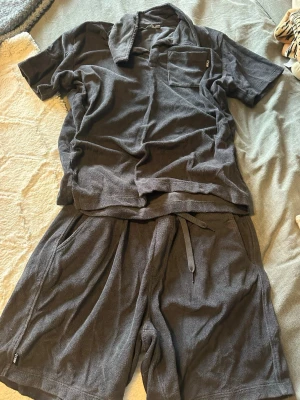 Mörkgrå frotté t-shirt från Warp - Säljer ett mörkgrått sett från Warp i mjuk frotté. T-shirten har korta ärmar, krage och en bröstficka med liten Warp-logga. Perfekt för chill dagar och har en loose passform. Materialet är riktigt skönt mot huden och ger en avslappnad vibe.