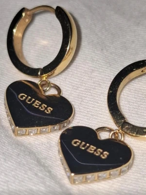 Örhängen Guess - Snygga örhängen från Guess med hjärtformad design i guld. Örhängen har guld detaljer med glittrande stenar längs kanterna. Perfekt för dig som gillar att sticka ut med stilrena accessoarer och vill ha något som känns lyxigt. Passar bra med allt.