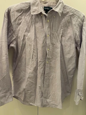 Ljusgrå skjorta från Ralph Lauren - Säljer en stilren ljusgrå skjorta från Ralph Lauren med klassisk krage och knappar hela vägen. Skjortan har lång ärm och den ikoniska broderade loggan på bröstet. Perfekt för dig som gillar enkel och snygg design.