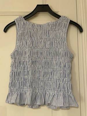Blåvit randig ärmlös linnetopp - Säljer då den är för stor för mig, använd en gång. Köpt på Plick som ny, men vet ej märket. Säljer en söt ärmlös linnetopp med blå och vita ränder. Toppen har smockad struktur och en volangkant längst ner som ger en extra touch. Perfekt för varma dagar och har en fräsch, somrig vibe. Materialet känns lätt och luftigt.