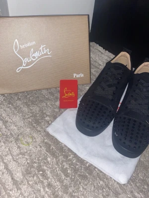 Christian Louboutin svarta sneakers med nitar - Säljer ett par svarta sneakers från Christian Louboutin med ikonisk röd sula och svarta nitar på tån. Skorna har snörning och är tillverkade i skinn med lyxig känsla. Perfekt för dig som vill sticka ut med exklusiv design och edgy detaljer.