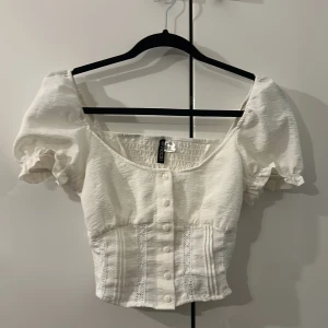 Vit blus med puffärm från H&M Divided - Säljer en vit blus från H&M Divided i storlek XS. Blusen har puffärmar, knappar framtill och fina spetsdetaljer på framsidan. Materialet är lätt och har en skrynklig struktur, med smock baktill för extra komfort.