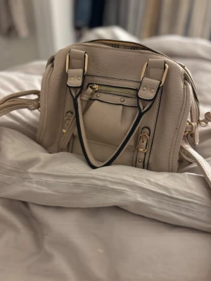 Beige axelväska med gulddetaljer - Säljer en beige axelväska i skinnimitation med guldfärgade metalldetaljer och dragkedja framtill. Väskan har både handtag och justerbar axelrem, samt dekorativa spännen och tofsar på sidorna. Perfekt storlek för det viktigaste och snygg design.