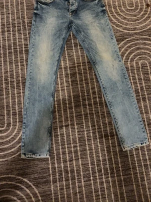 Ljusblå jeans från Denim Rivalry - Säljer ett par ljusblå jeans från Denim Rivalry med snyggt slitna detaljer och klassisk femficksdesign. Jeansen har raka ben och normal passform, tillverkade i mjukt bomullsmaterial. Perfekt för dig som gillar en chill och avslappnad stil.