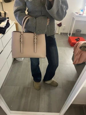 Ljus rosa handväska med axelrem - Säljer en stilren beige/rosa handväska med både handtag och justerbar axelrem. Väskan har guldfärgade detaljer och flera fack inuti, inklusive ett med dragkedja. Materialet är slätt och känns som syntet/skinn. Perfekt storlek för att få plats med allt du behöver. 💕