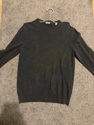 Mörkgrå stickad tröja från Esprit - Snygg mörkgrå stickad tröja från Esprit med diskret logga på bröstet. Tröjan har rund halsringning och långa ärmar. Materialet är mjukt och känns skönt mot huden. Passar dig som gillar enkel och stilren design.