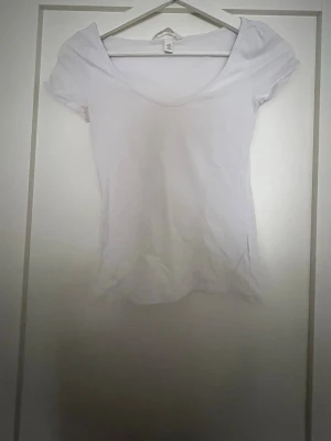 Vit basic topp med rund hals - En enkel vit topp med rundad, djup urringning och korta ärmar. Modellen är figurnära och passar perfekt till jeans eller kjol. Tillverkad i mjuk bomull som känns skön mot huden. Perfekt för en clean och fräsch look.