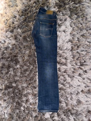 Blå jeans från Nudie Jeans Co - Snygga blå jeans från Nudie Jeans Co med klassisk femficksdesign och orange kontrastsömmar. Jeansen har en rak passform och är tillverkade i slitstarkt denimtyg. Perfekta för dig som gillar en tidlös och stilren look.