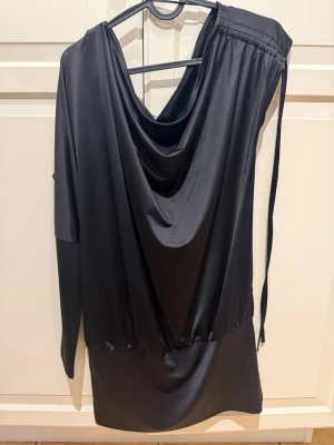 Svart en-ärmad med drapering - Snygg svart kortklänning från Monki med draperad front. Klänningen har lång ärm på ena sidan och ärmlös på andra. Tillverkad i mjukt, glansigt syntetmaterial och har knytband vid axeln.