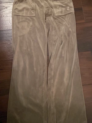 Gröna mjukisbyxor Juicy Couture XS - Säljer ett par gröna mjukisbyxor från Juicy Couture i storlek XS. Byxorna har breda ben, mjukt sammetstyg och stora fickor med guldknappar framtill. Klassisk Juicy Couture-logga broderad på ena fickan. Perfekta för chill och comfy vibes.