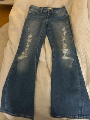 Blå bootcut jeans med slitningar från Gina Tricot - Säljer ett par blå bootcut jeans med snygga slitningar framtill och detaljerade bakfickor med knapp. Jeansen har låg midja och är tillverkade i klassiskt denim-material. 